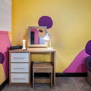 A small wooden desk with a mirror, a desk lamp, and a drawer unit against a yellow wall with purple accents. - Social Media Adventure - 1 Bed 10% off long stays (Doncaster)