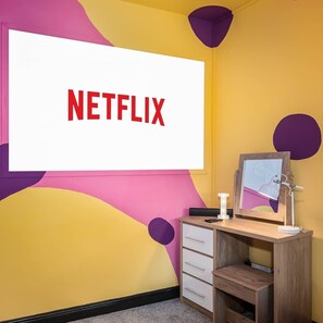 A room with a large TV screen displaying "NETFLIX" and a wooden desk with a mirror, drawers, and a lamp.