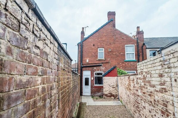 A narrow alleyway with a brick wall on one side and a red brick house with a white door and windows on the other.
