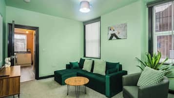 A living room with a green sofa set, a wooden coffee table, and a green armchair. There is a painting on the wall and a potted plant by the window.