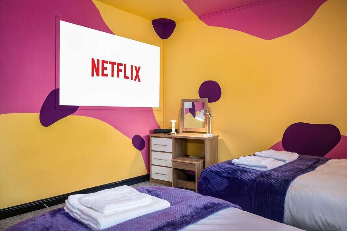 Ein Hotelzimmer mit einem Bett, einem Schreibtisch mit Spiegel und einem an der Wand befestigten Bildschirm, der das Netflix-Logo zeigt.