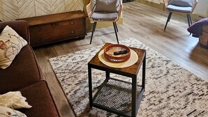 A living room with a herringbone-patterned wall, a brown sofa, a wicker chair, a small wooden table with a woven coaster, and a patterned rug.