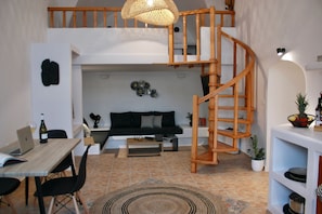 A modern loft apartment with a wooden spiral staircase, a black sofa, a dining area with a laptop and wine bottle, and a small kitchen area. - The Lithos Villa | The Perfect retreat to enjoy the immense beauty of Santorini (Thira)