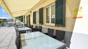 Un café en plein air avec des tables et des chaises sous un auvent, un panneau indiquant les consignes de sécurité pour la piscine, et une vue sur la mer.