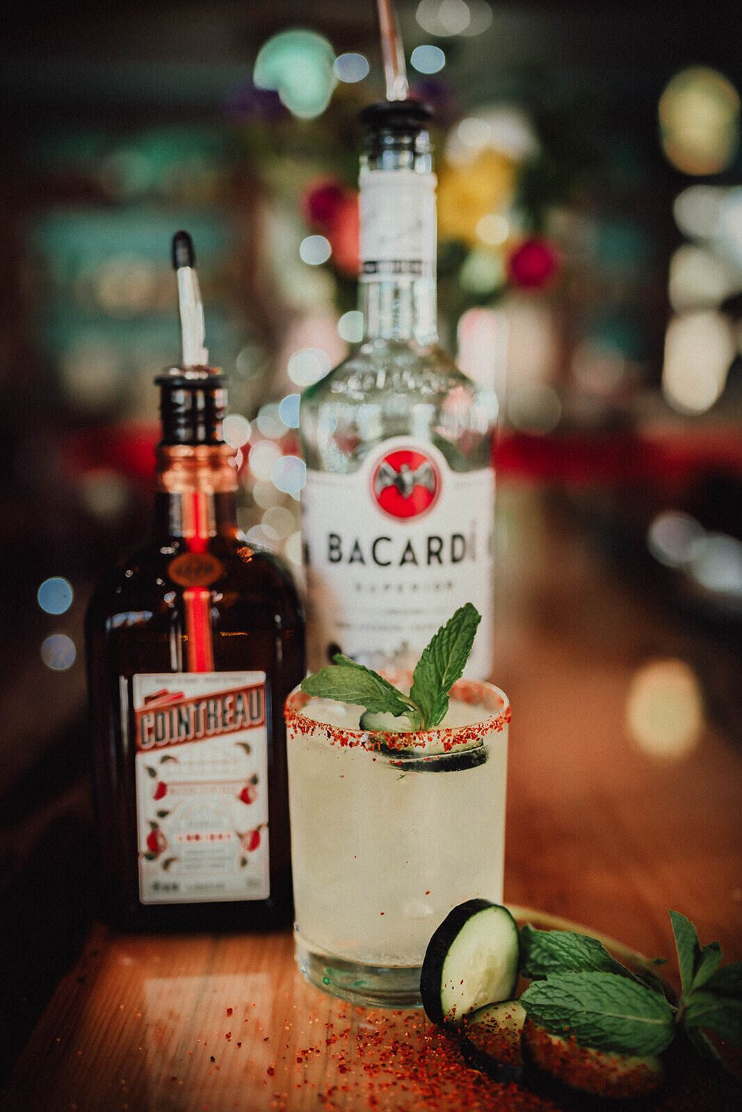 Un bar con una botella de Cointreau y una de Bacardí, un cóctel adornado con pepino y especias rojas esparcidas sobre la barra.