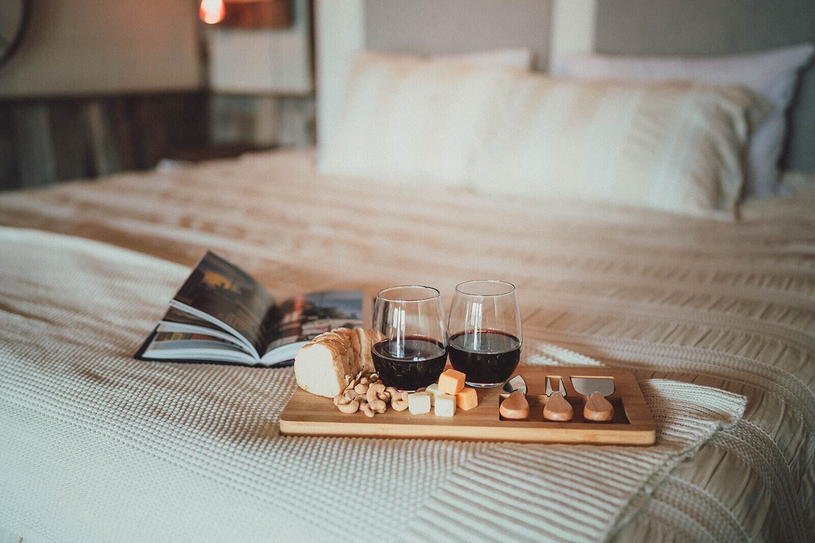 A bed with a wooden tray holding a book, two glasses of red wine, and cheese.