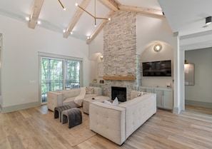 A spacious living room with a stone fireplace, a large window, and a flat-screen TV mounted on the wall. - New Lakeside Home w/kayaks, Game Room, Billiards (Winchester)