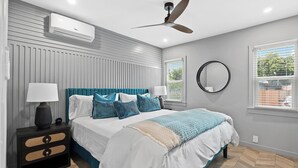 Ein modernes Schlafzimmer mit Bett, Nachttischen, Deckenventilator, Spiegel und Fenster mit Jalousien.