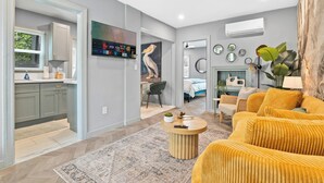 Ein modernes Wohnzimmer mit einer Couch, einem Couchtisch und einem Fernseher. Es gibt eine Küchenzeile mit Schränken und einem Fenster.