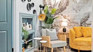 Ein gemütliches Wohnzimmer mit einer blauen Tür, einem gelben Sofa, einem grauen Sessel und einer großen Pflanze.