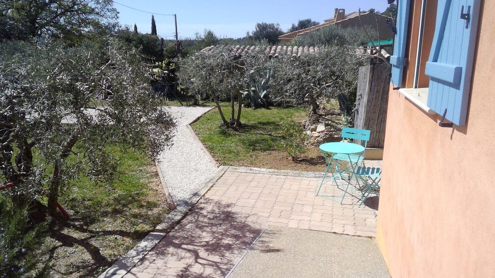 A paved pathway leading to a small outdoor seating area with a table and chairs, surrounded by olive trees and a building with blue shutters.