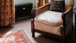 Une pièce avec une chaise en bois, un radiateur, une fenêtre ornée de rideaux à motifs floraux et une lampe.