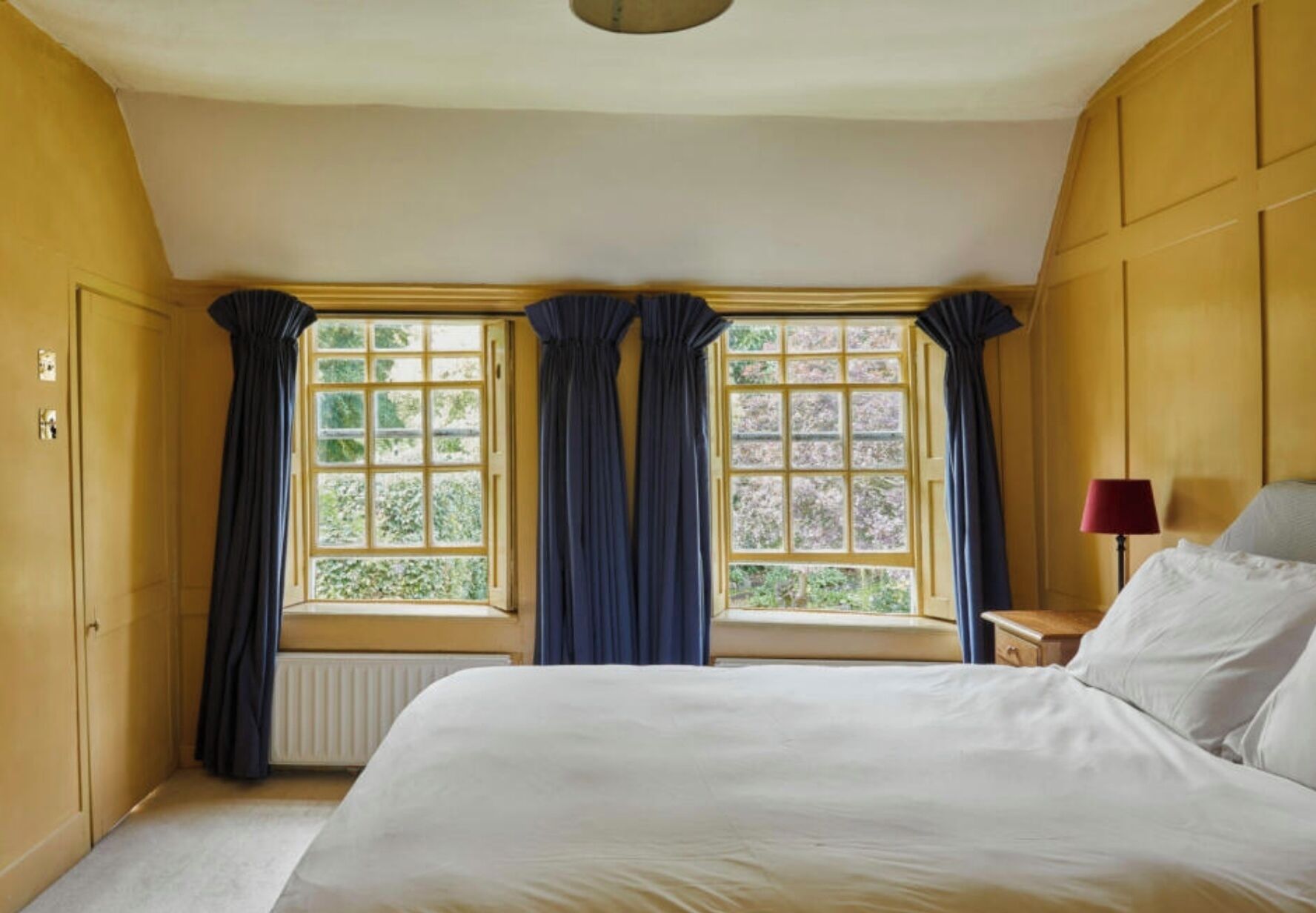 A bedroom with a large bed, two windows with curtains, a radiator, and a bedside table with a lamp.