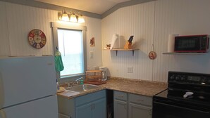 Una cocina equipada con refrigerador, pileta, cocina y microondas. Se aprecia una ventana con persianas, un reloj y un adorno en la pared.