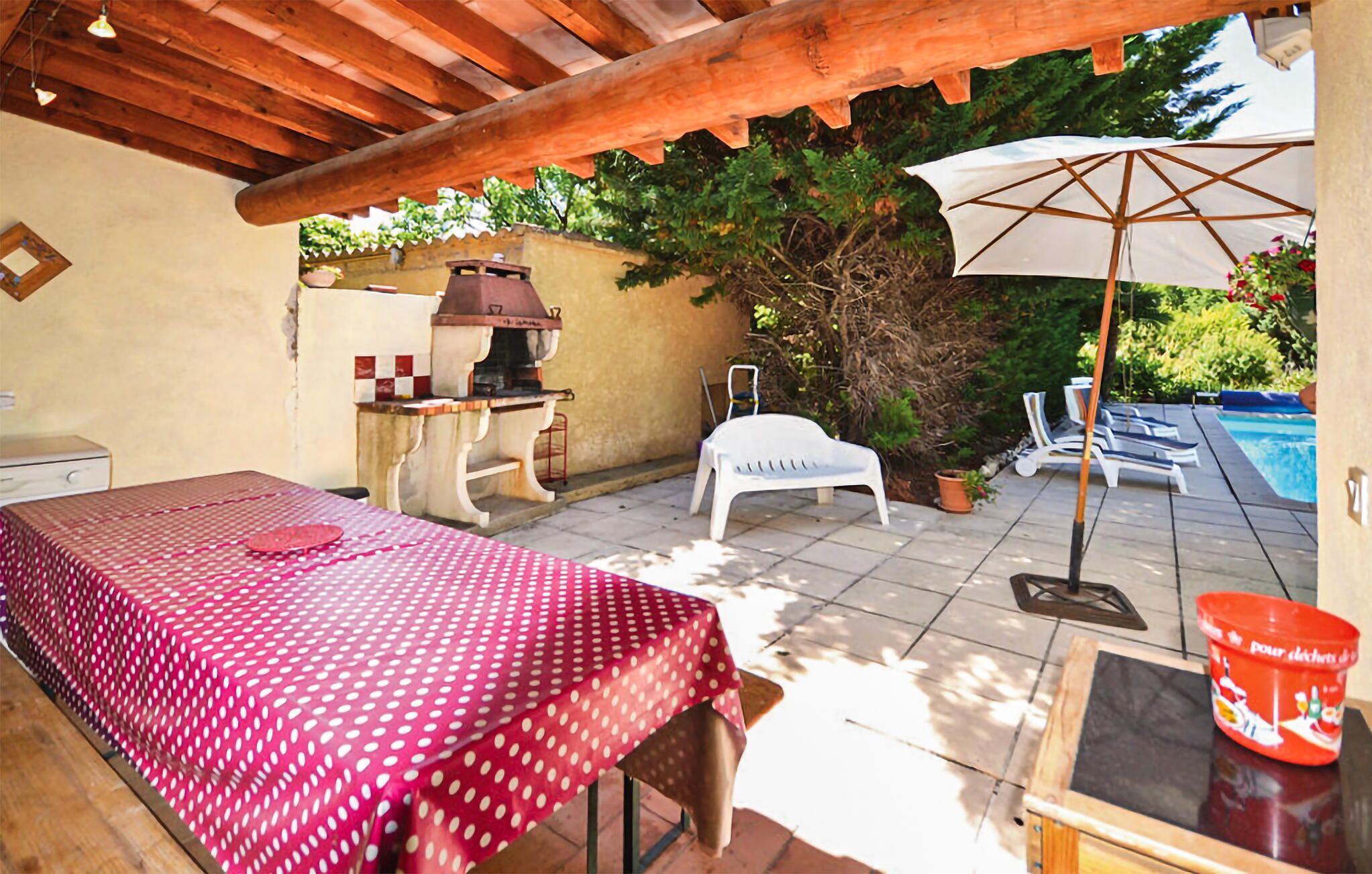 Une terrasse extérieure avec une table recouverte d’une nappe à pois rouges, un barbecue, un parasol blanc et un espace piscine avec des chaises longues.