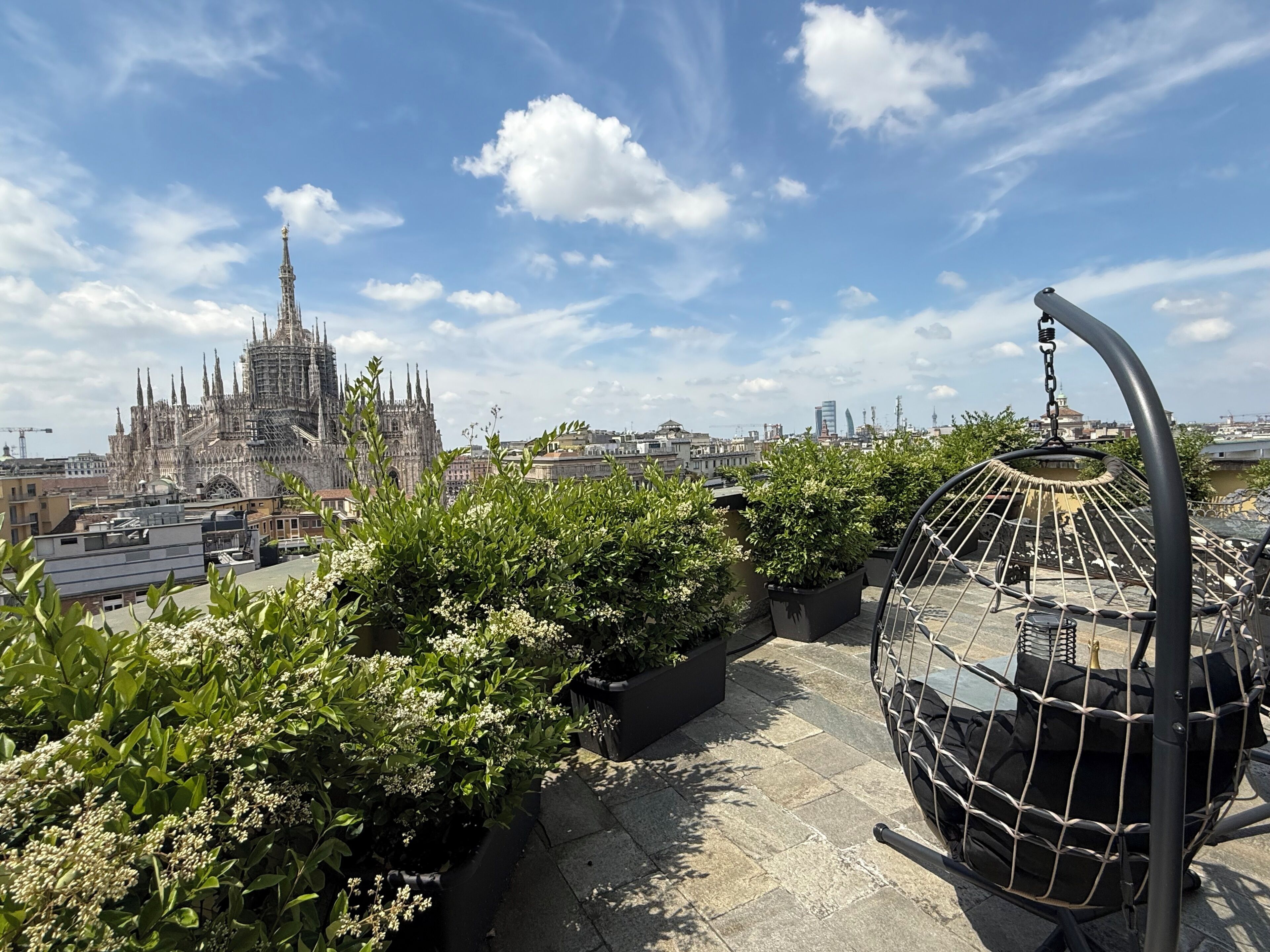 A rooftop garden with potted plants and a hanging chair, overlooking a historic cathedral and cityscape.