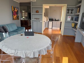 A modern living room with a round table, clear chairs, a sofa, and a kitchen in the background.