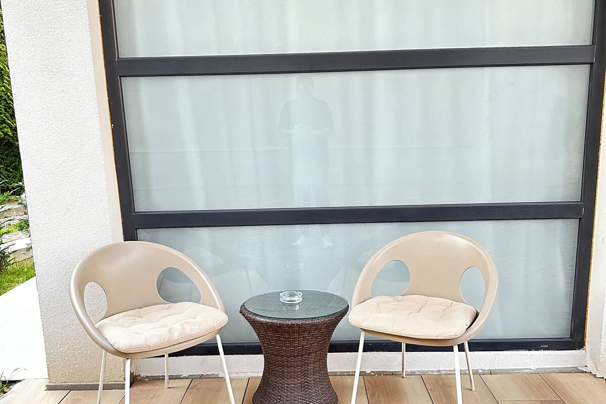 Un patio con dos sillas color beige y una pequeña mesa redonda, frente a un ventanal con marco negro.
