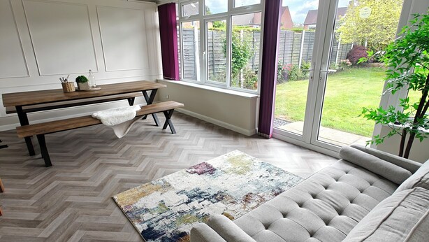 Ein modernes Wohnzimmer mit Fischgrätparkett, einem grauen Sofa, einer Holzbank und einem großen Fenster mit Blick auf einen Garten.