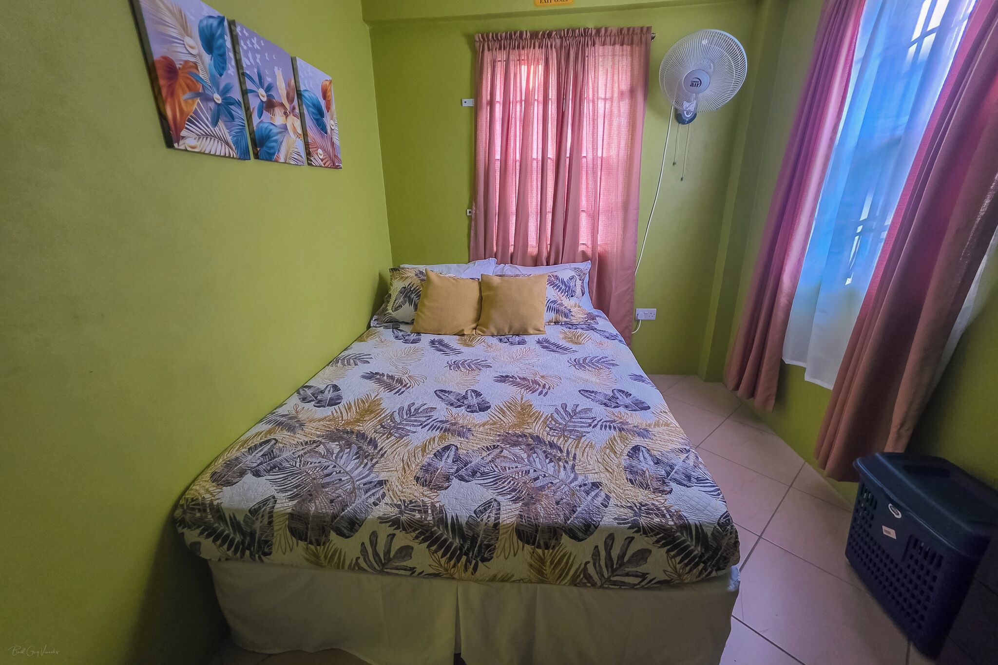 A room with a bed, a fan, curtains, and a wall with artwork.