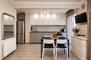 A modern kitchen with white cabinets, a dining table with two chairs, a refrigerator, and a flat-screen TV mounted on the wall.