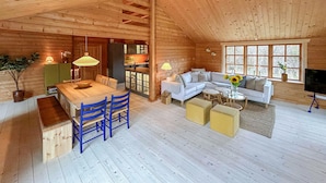 Een gezellig houten blokhut interieur met een eethoek, een woonkamer met een bank en salontafel, en een keuken zichtbaar door een open deur.