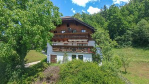 A two-story wooden chalet with a balcony, surrounded by lush greenery and trees.