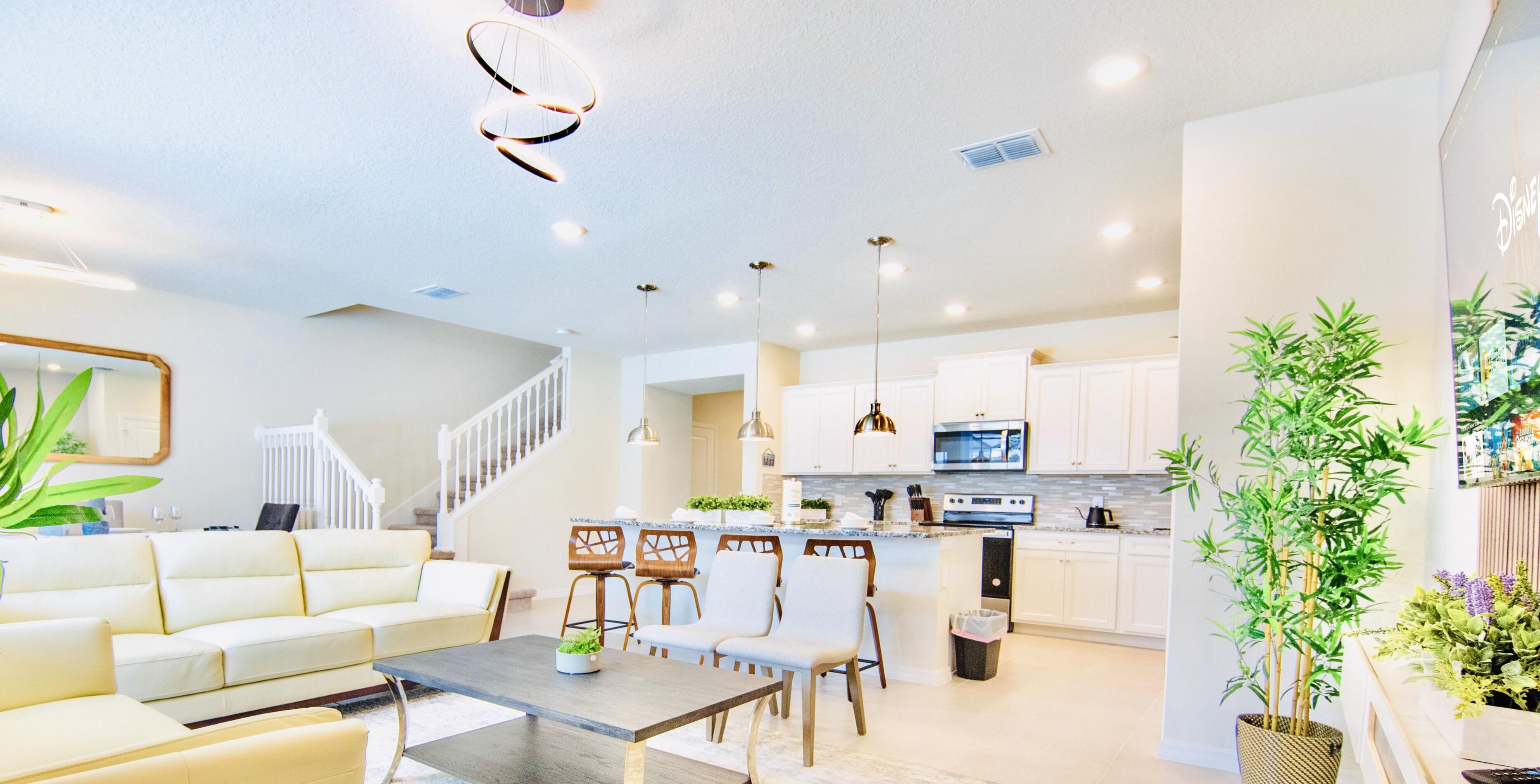 A modern living room with a sofa, dining table, and chairs, a kitchen with white cabinets and appliances, and a staircase leading to another level.