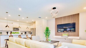 Un salon moderne avec un canapé blanc, un coin repas avec des chaises, une cuisine avec des meubles blancs et un téléviseur fixé au mur diffusant du contenu Disney.