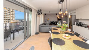 A modern kitchen with a dining area, a balcony with a view of the sea, and a living area with a sofa and TV.