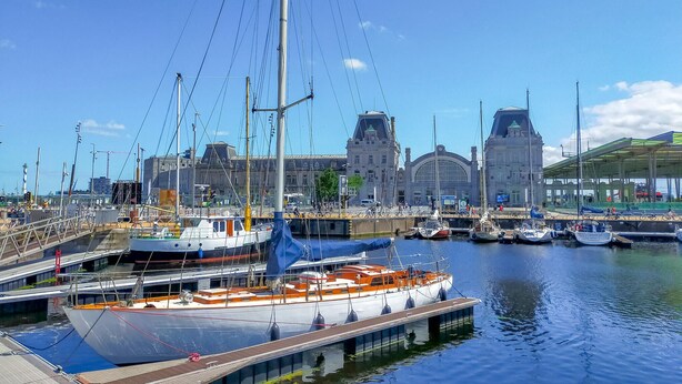 A marina with boats docked, a large building with a clock tower in the background, and a covered area with bicycles.