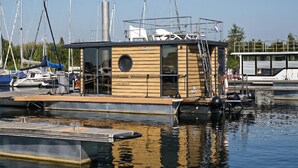 A wooden floating structure with a circular window, surrounded by calm water and docked boats. - Stunning ship in Ophoven with WiFi (Ophoven)