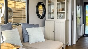 A living room with a beige sofa, white and blue cushions, a white cabinet with glass doors, and a round mirror on the wall. - Stunning ship in Ophoven with WiFi (Ophoven)