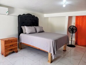 Une chambre à coucher avec un lit, une table de chevet en bois, un ventilateur sur pied et un sol carrelé.