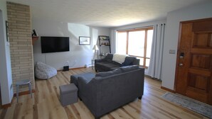 A living room with a grey sofa, a small table, and a television mounted on the wall. - The Maine House in State College - 2 miles to PSU via walking path or car! (State College)