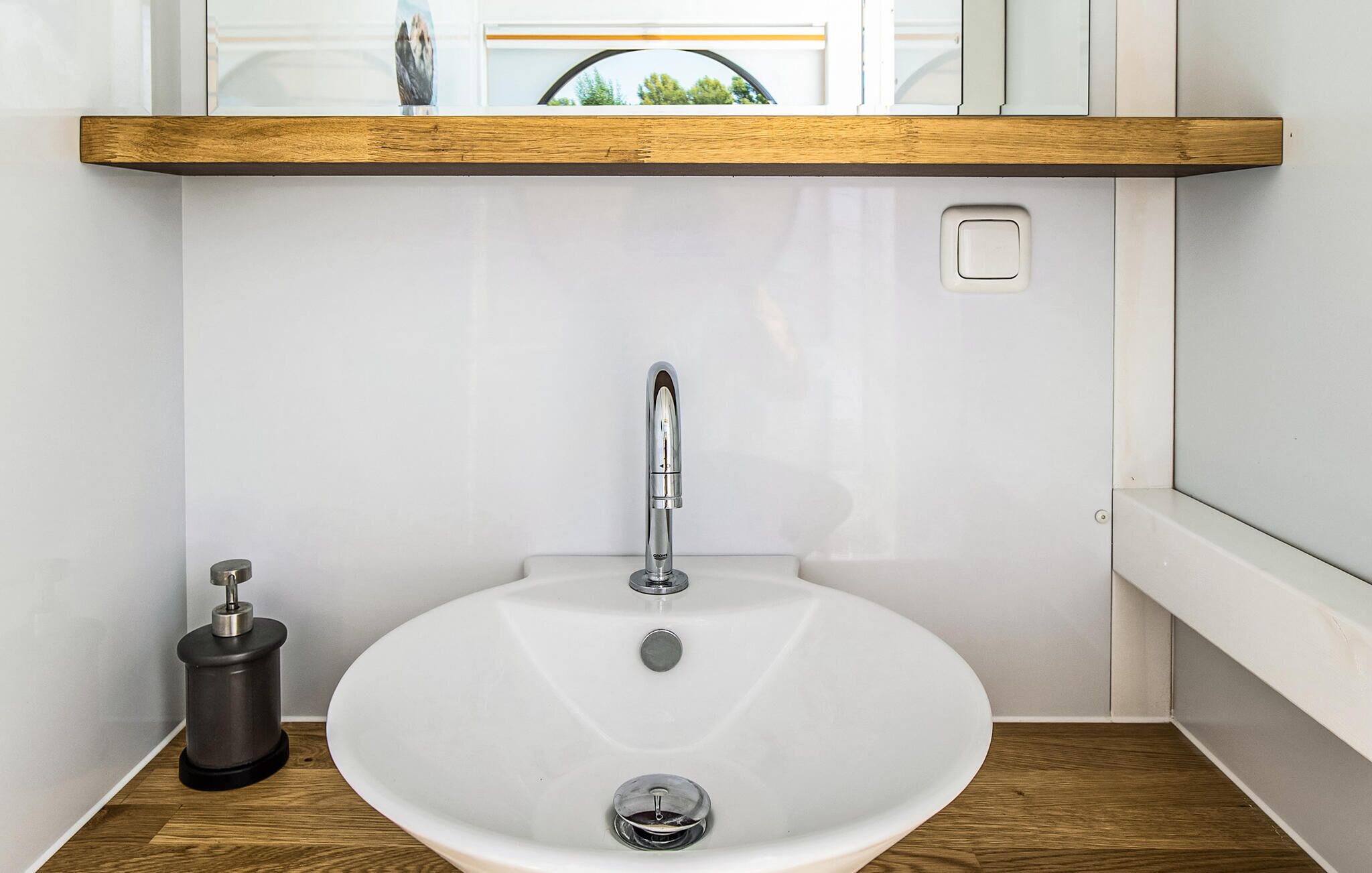 A bathroom with a white sink, a modern faucet, a wooden shelf above the sink, and a trash can.