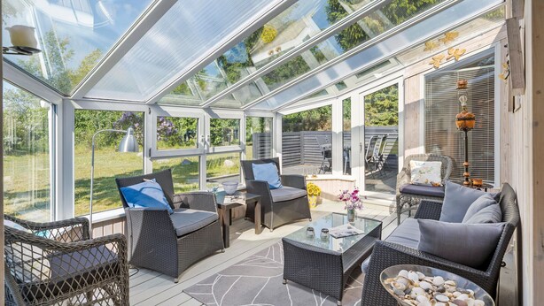 Une véranda meublée de mobilier en osier, dotée de tables en verre et offrant une vue sur le jardin.