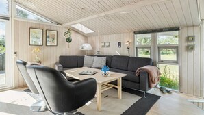 Un salon moderne avec un plafond en bois, un canapé gris, un fauteuil noir et une table en bois. Il y a deux fenêtres donnant sur un paysage verdoyant.