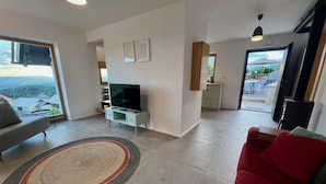 Una sala de estar moderna con un televisor de pantalla plana sobre un mueble blanco, un sofá rojo, un sofá gris y un ventanal con vistas al exterior.