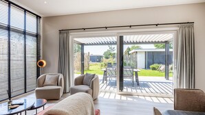 Ein modernes Wohnzimmer mit einer Couch, Sesseln und einem Couchtisch. Eine Glastür führt auf eine Terrassenförmige Außenfläche mit Tisch und Stühlen.