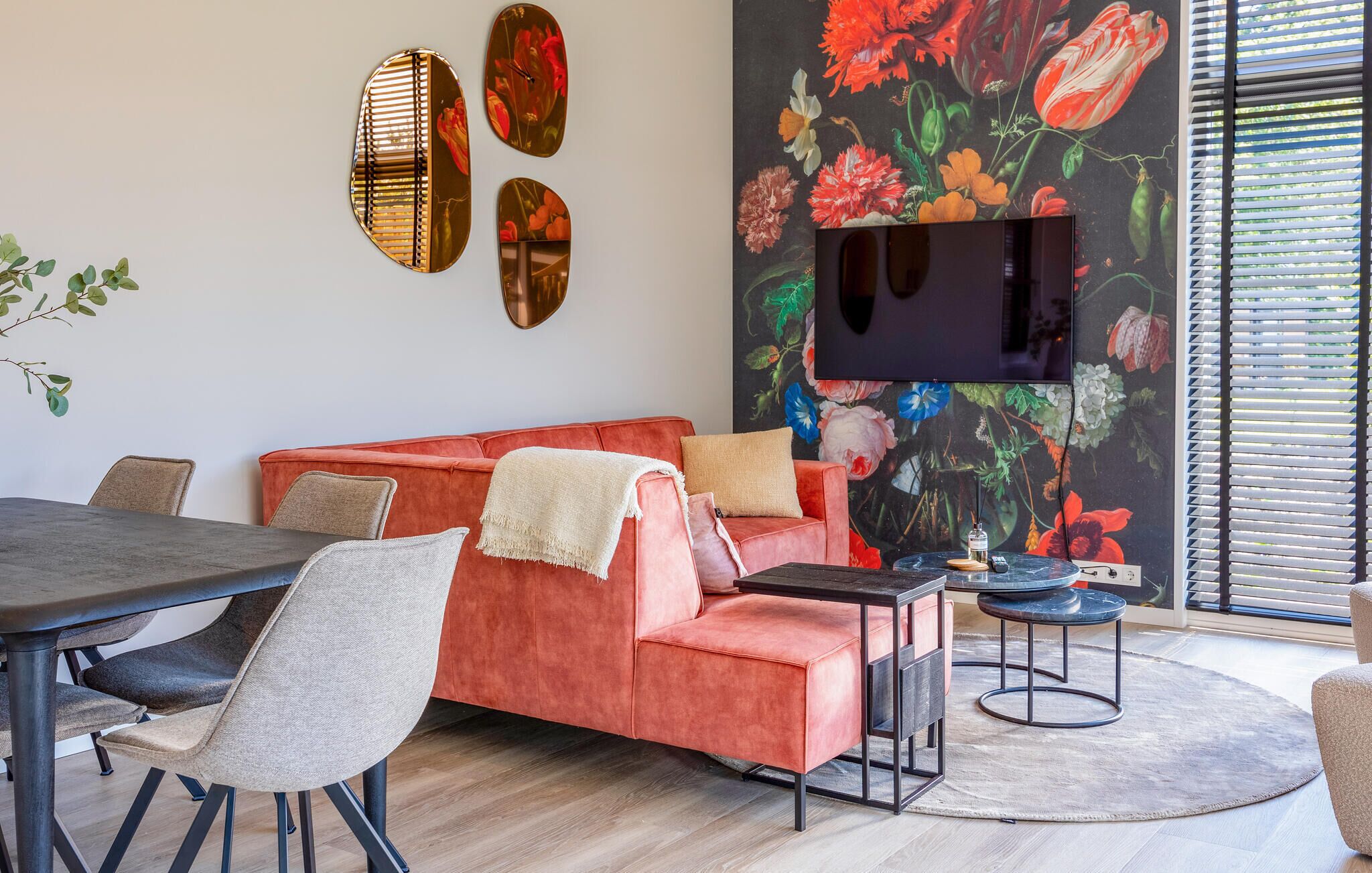Un salon moderne avec un canapé rouge, une table à manger et des chaises. Un téléviseur à écran plat est fixé au mur, et il y a une grande fresque murale à motifs floraux.