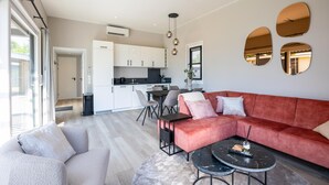 Ein modernes Wohnzimmer mit einem roten Sofa, grauen Sesseln und einem Couchtisch. Im Hintergrund ist eine Küche mit weißen Schränken und einem Essbereich zu sehen.