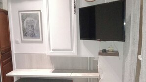 Une pièce avec un bureau blanc, un petit tabouret, un tableau encadré, une horloge et un téléviseur à écran plat fixé au mur.