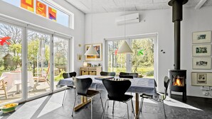 A modern living room with a large fireplace, a dining area with a table and chairs, and large windows offering a view of the outdoors.