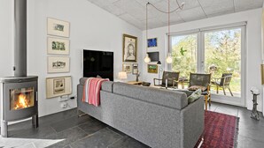 A modern living room with a stone fireplace, a grey sofa, a TV, and a large window with a view of trees.