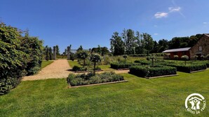 Un jardin bien entretenu, avec un chemin de gravier menant à un bâtiment en pierre, entouré de haies et d’arbres soigneusement taillés.