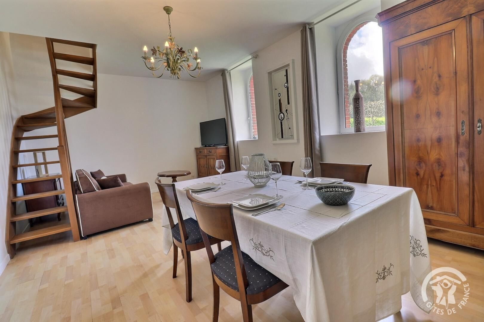 Uma sala de jantar com mesa posta para quatro pessoas, um candelabro, uma escadaria de madeira e uma grande janela em arco.