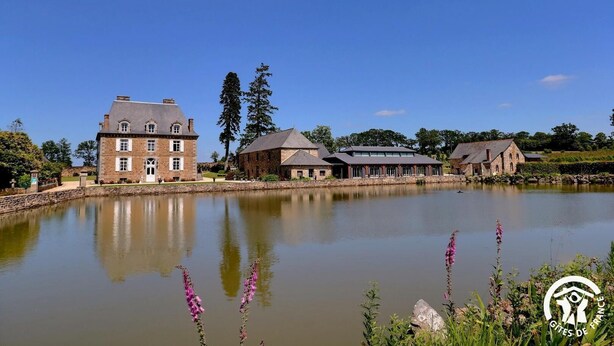 Un grand bâtiment en pierre, avec un étang devant, entouré d’arbres et sous un ciel dégagé.