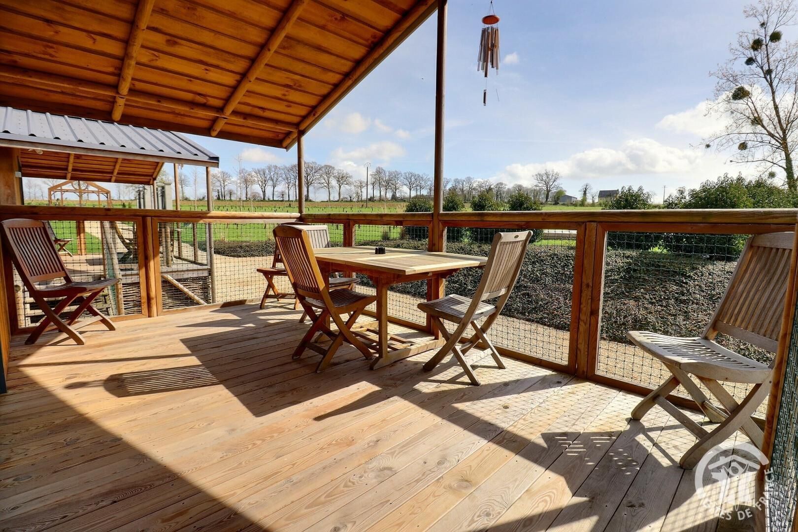A wooden deck with a table and four chairs, overlooking a fenced area with trees and a clear sky.