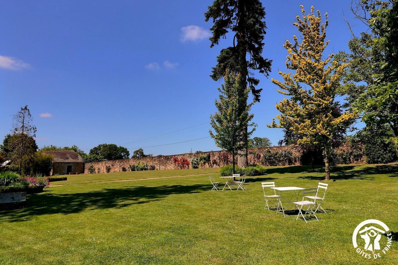 An outdoor seating area with tables and chairs, a stone wall, and trees in the background.